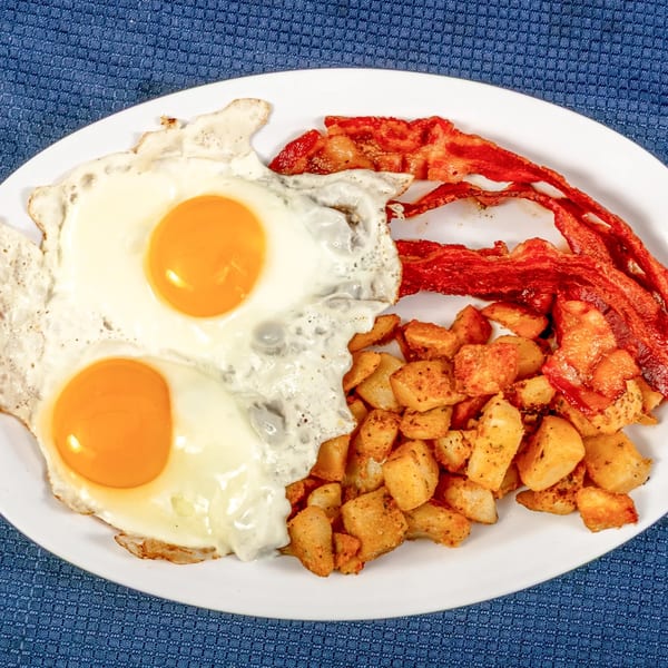 A diner breakfast with two fried eggs, crispy bacon, and seasoned home fries on a white plate.